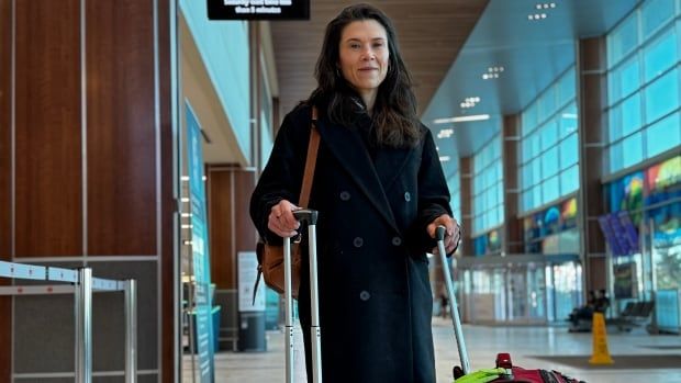 A white woman is wearing a long, black coat with hands on her the handles of two suitcases.