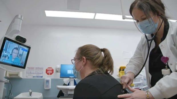 A nurse uses an electronic stethoscope to listen to a patient's lungs, while a remote doctor, pictured on a screen in front of the patient, watches and listens to a live-feed.