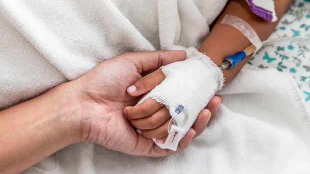 An adult holds a child's hand that has an IV in it. The hands are against a white blanket.