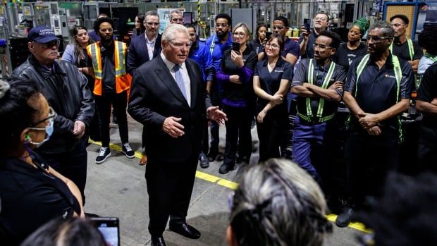 A man in a suit stands in a factory setting with workers around him looking at him as he speaks