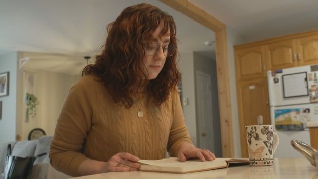 A woman with wavy red hair sits at a kitchen counter looking over an open book. There's a butterfly mug next to her. 