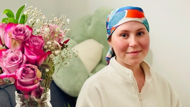 A young woman with a colourful headwrap smiles at the camera. She is wearing a white sweater and sitting on a couch with a fuzzy stuffed animal behind her. There are roses to her left