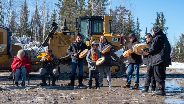 A group of people are seen using hand drums at a muddy construction site.