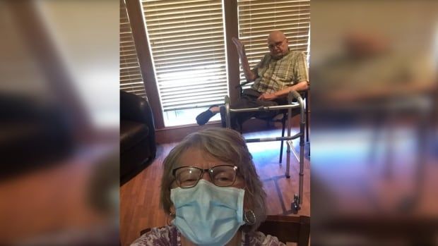 A woman in a face mask takes a selfie with a man sitting in a chair behind her.