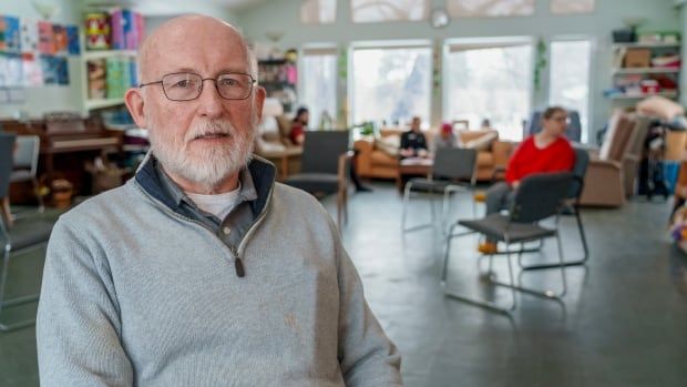 A bald man with a grey beard wears a grey sweater and looks into the camera. He wears a concerned look on his face. 