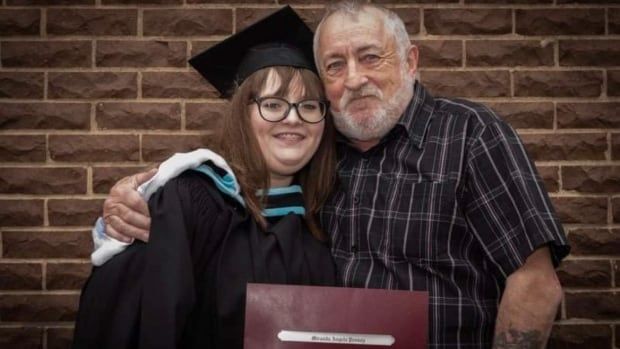 A woman (left) stands with her dad(right) holding ger graduation certificate at a ceremony.