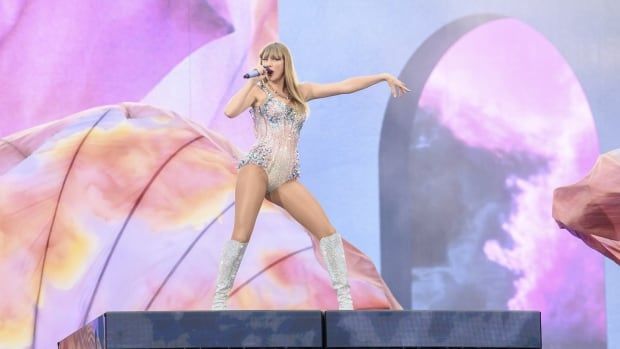 A blonde-haired woman poses with a microphone on a stage against a colourful pastel orange, pink and blue backdrop, wearing a sparkly bodysuit. 