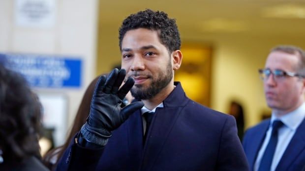 A man wearing a blue jacket and black gloves smiles and waves to the camera.