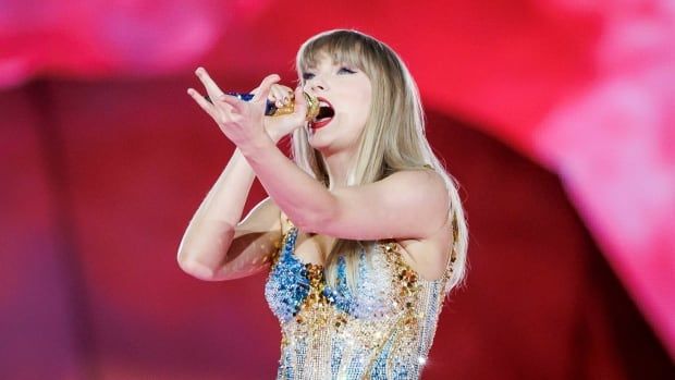 A woman in a sequined blue-tinged gown stands at a microphone in front of a red backdrop with a hand raised palm-up in front of her as she belts out a song.