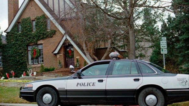 An older model of police car sits outside of a brown house with triangular peaks over the front door and the overall shape of the house. 