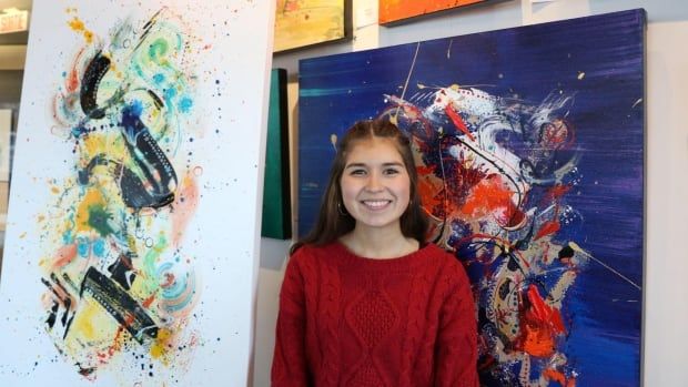 A teen smiles, standing in front of several abstract paintings hung on the wall. 