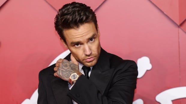 A dark haired man in a suit raises his tattooed hand in a red carpet photo.