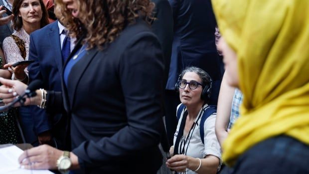 A woman holding a podcast recording device is sitting amongst a court hearing, recording what is happening. 