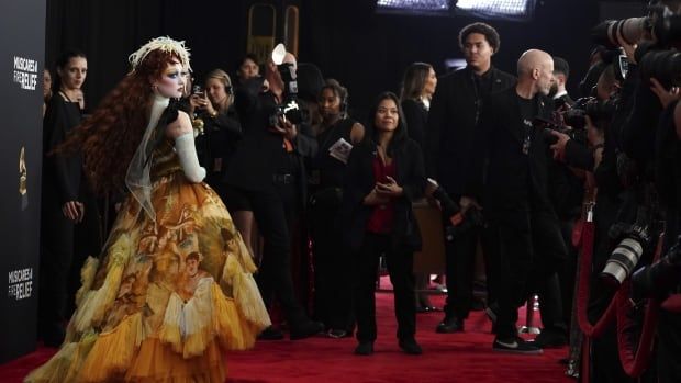 A woman in a feathery, layered yellow dress is pictured on a red carpet with a number of photographers visible beyond her. 