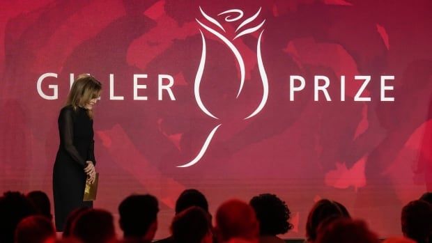 A woman stands onstage in front of a red backdrop that says Giller Prize with a stylized rose drawing in the centre. At the bottom of the image, the heads of a crowd are visible. 