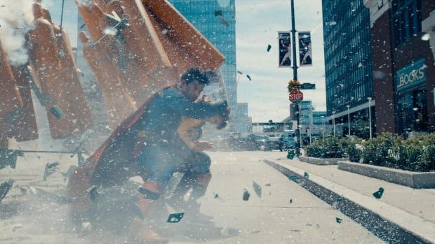 Small flecks of debris fill the screen as Superman protects a small child from an unknown threat on a city street. 