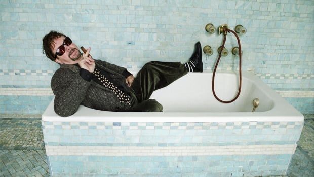 A man wearing a suit and smoking a cigar lies back in a bathtub.