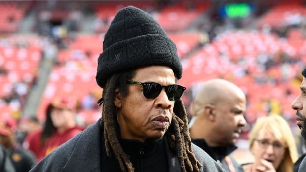 A man with dreadlocks wearing sunglasses, a dark knit hat and a grey coat stands among others on the field in a football stadium. 