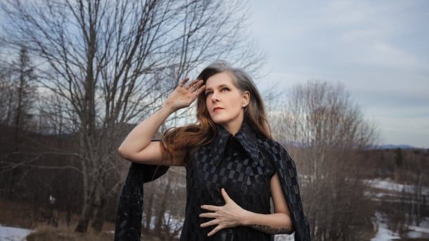 A woman stands on a snowy hill, one hand on her stomach, the other brushing her hair from her brow.