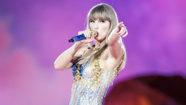 On stage in a sequinned bodice suit, Taylor Swift with mic in one hand, and pointing to the audience with her other hand.