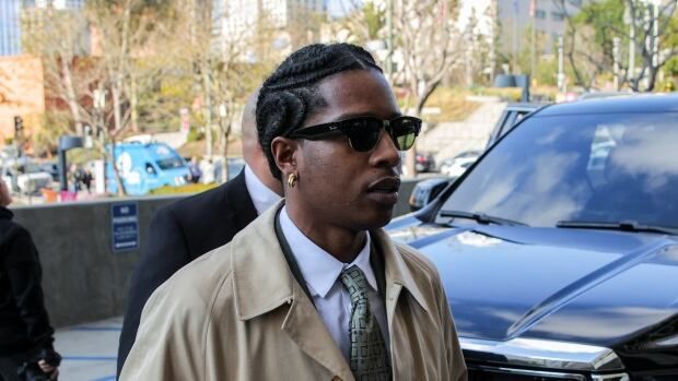 A man with dark sunglasses and a beige trenchcoat over a suit and tie is seen in front of a black SUV.