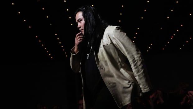 An Indigenous musician with long dark hair steps on stage to accept an award. The background is mostly black with some lights.