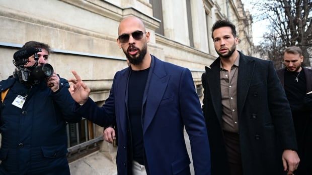 Two men with beards, one bald, walk on a sidewalk as media and photographers surround them.