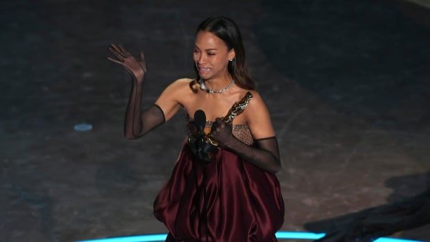 A woman in a burgundy dress cries on stage as she holds a gold statuette.