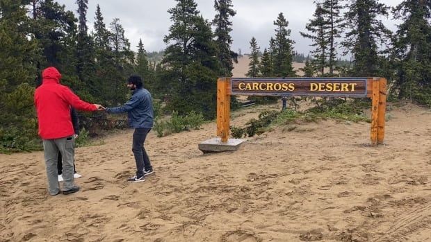 Two men stand in the sand near a sign reading, 'Carcross Desert.'