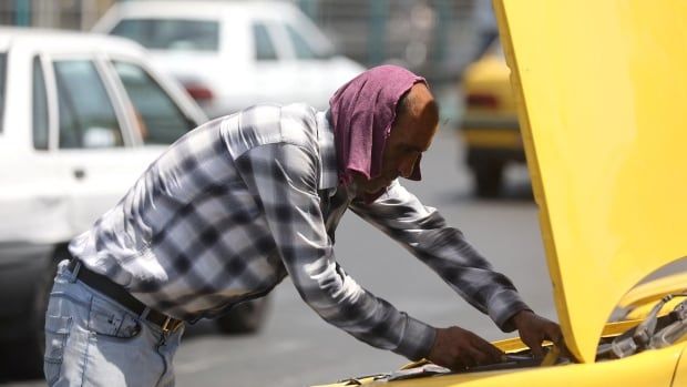 A man with a shirt over his head looks down at the hood of a car.