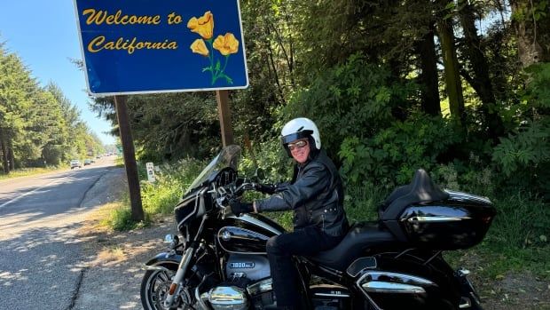 Bob McDonald sits on his black sporty motorcycle with his white helmet in the shade on the side of the road under a sign that says, "Welcome to California."