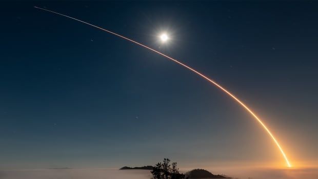 A long exposure photo of a rocket blasting up at night shows a curved orange streak against the sky with the sun rising off in the distance. 