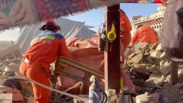 A still from video shows people in helmets and orange suits sifting through rubble and concrete debris.