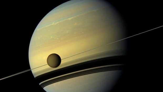 Saturn is yellow with brown stripes. Rings surround it with a small moon in the front in front of the planet.