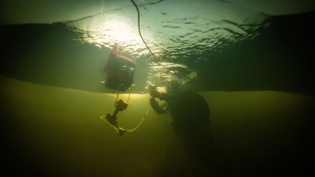 a man in scuba gear under the water is surrounded by tubes and technical equipment. The waters surface is covered with ice, making the water around him look green and eerie