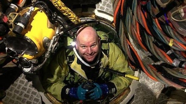 A man in a yellow diving suit pokes his head and shoulders up through a piece of machinery