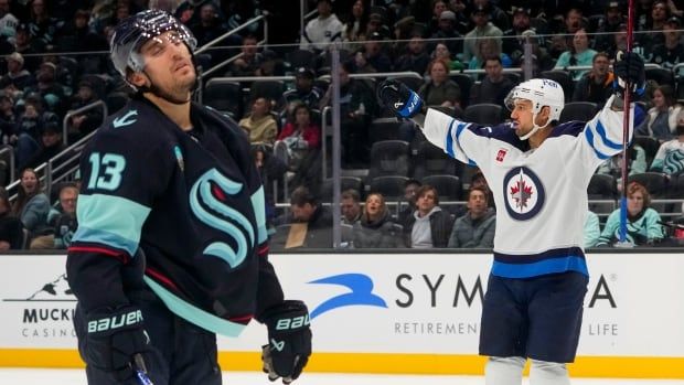 A hockey player in white and blue raises his arms in the air in celebration, while an opposing player in teal and navy reacts disappointedly.