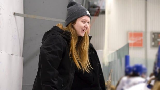 A smiling woman in a tuque stands behind hockey players sitting on a bench.