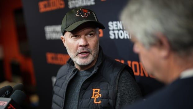 A man wearing BC Lions gear speaks near a bunch of microphones