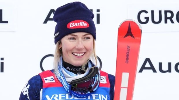 A women's skier is shown after a race, wearing a winter cap, as she poses on the podium while holding her skis.