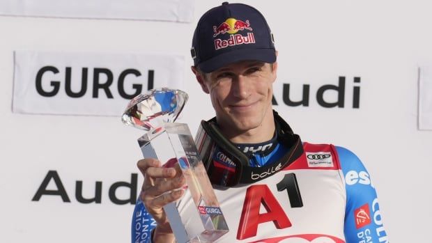 A men's skier holds a trophy and smiles while posing for a photo on the podium.