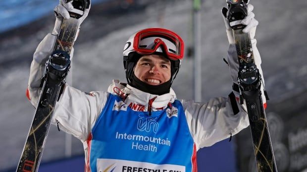 A male skier smiles whiles raising a ski in both hands.