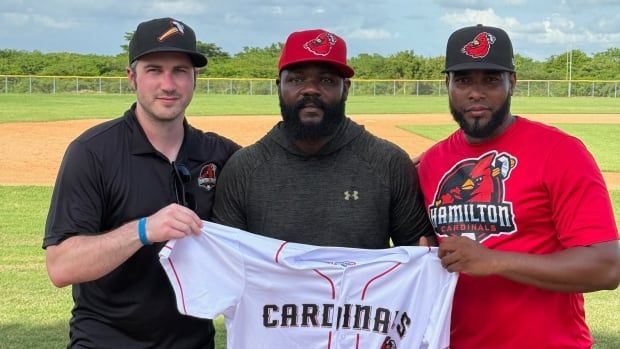 A Black man with a baseball cap holds up a Cardinals jersey with two men on either side of him. 