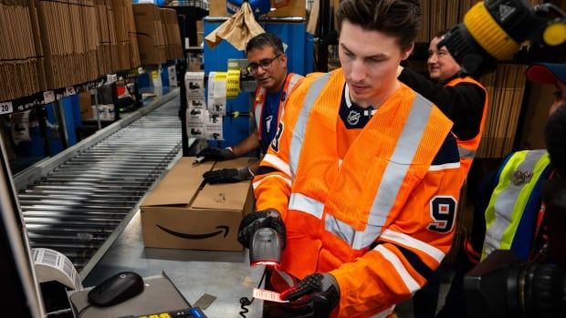 Hockey player in an orange vest stands next to a conveyer bet scanning items while two other men look on approvingly from the shipping giant.