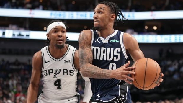 A male basketball player drives to the basket with both hands on the ball as an opposing player pursues him.