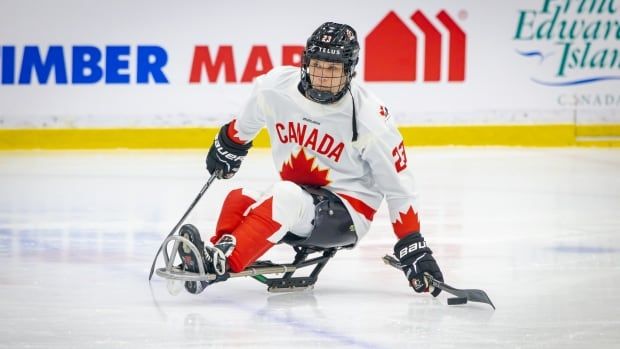 A man, wearing white and red hockey gear and a jersey that says "Canada" and a red maple leaf on it, is playing para hockey and is sitting on a double-blade sledge.