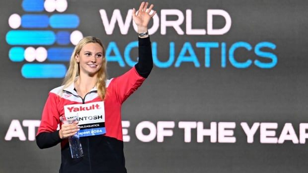 A female swimmer wearing a track suit smiles while waving with her right hand.