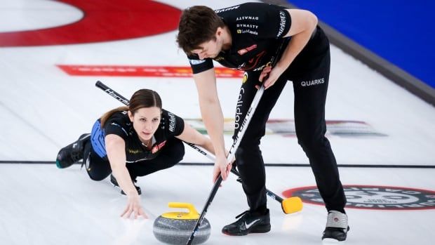 A female mixed doubles curling skip delivers a stone while her male partner sweeps.