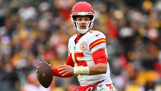 A male football quarterback looks to pass while holding the ball in his right hand during a game inside a stadium filled with fans.