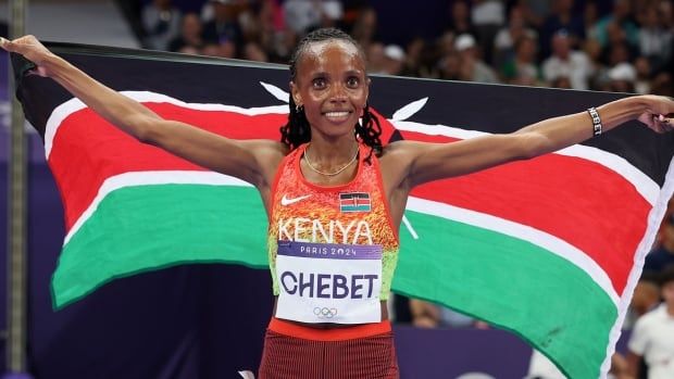 A female track athlete smiles while holding up the Kenyan flag behind her back.